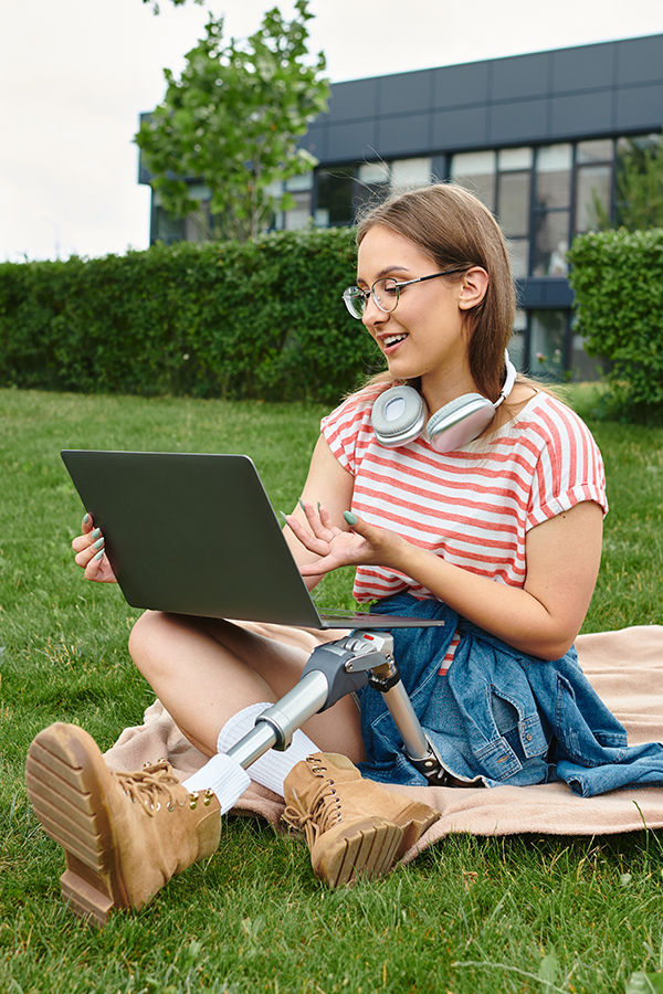 O tânără cu un picior protetic stă pe o pătură în parc, folosind un laptop. Ea zâmbește și poartă căști în jurul gâtului, având alături o cafea și un smartphone.